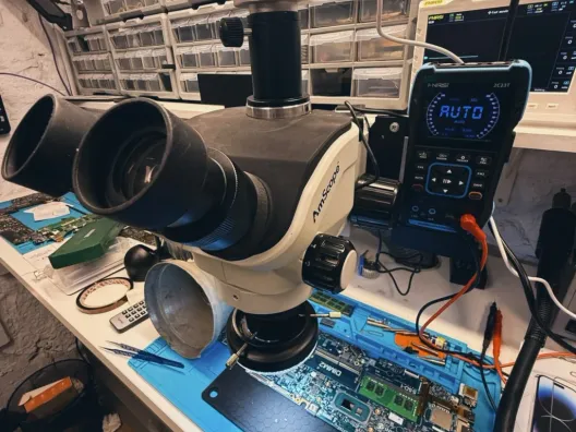 Banco de trabajo de BestFixing: microscopio estereoscópico, osciloscopio de mano y una placa de portátil PC con su módulo de memoria sobre el tapete antiestático