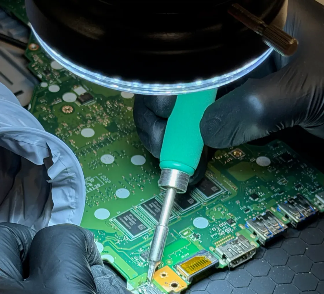 Manos con guantes de nitrilo trabajando con el soldador sobre la placa base verde de un portátil, bajo el anillo de luz