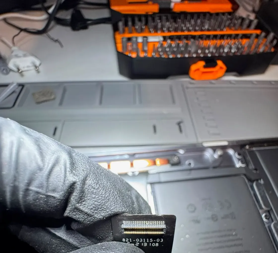 Mano con guante sujetando un cable plano de un portátil Apple con la referencia 821-03115-03 impresa, con el equipo abierto y la batería a la vista al fondo