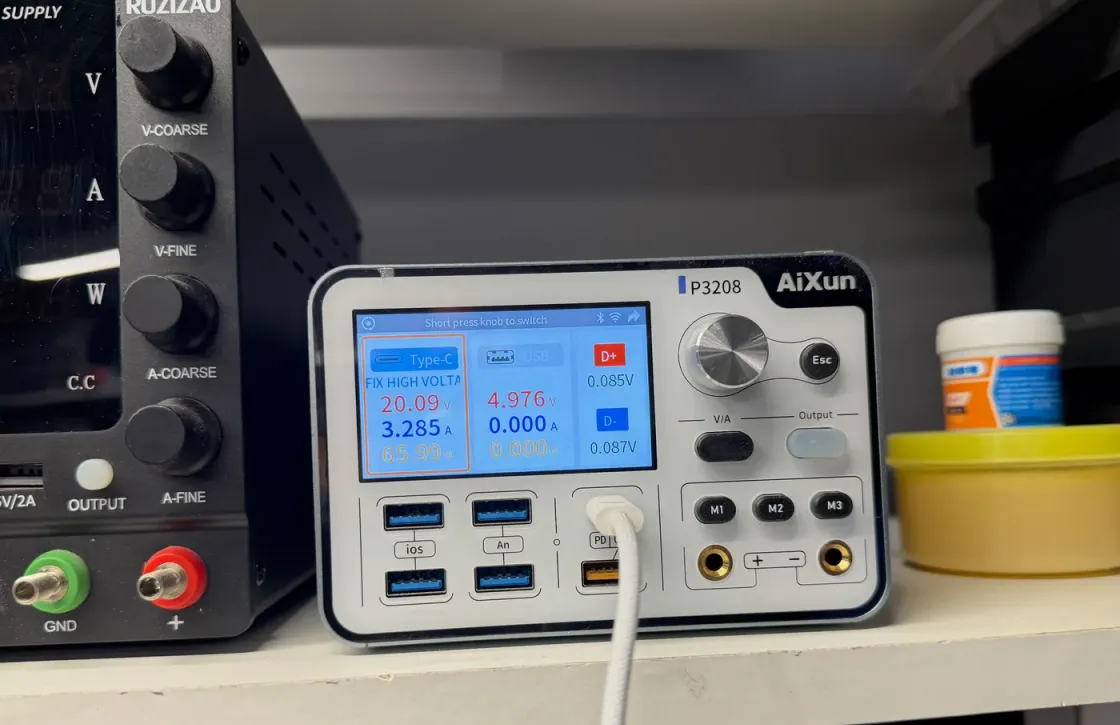 Fuente de alimentación de banco alimentando una placa en ensayo de carga: la pantalla marca 20,09 V y 4,976 A